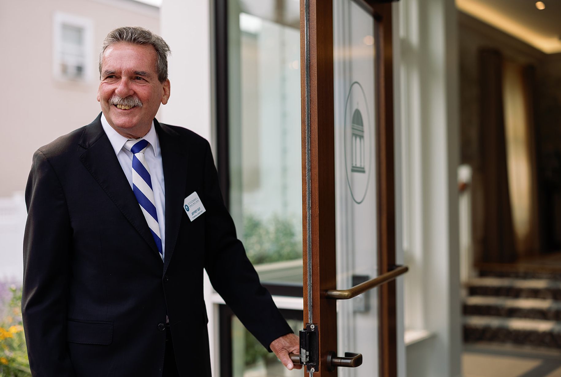 Man in suit opens a glass door, smiling. Building exterior, sunny.