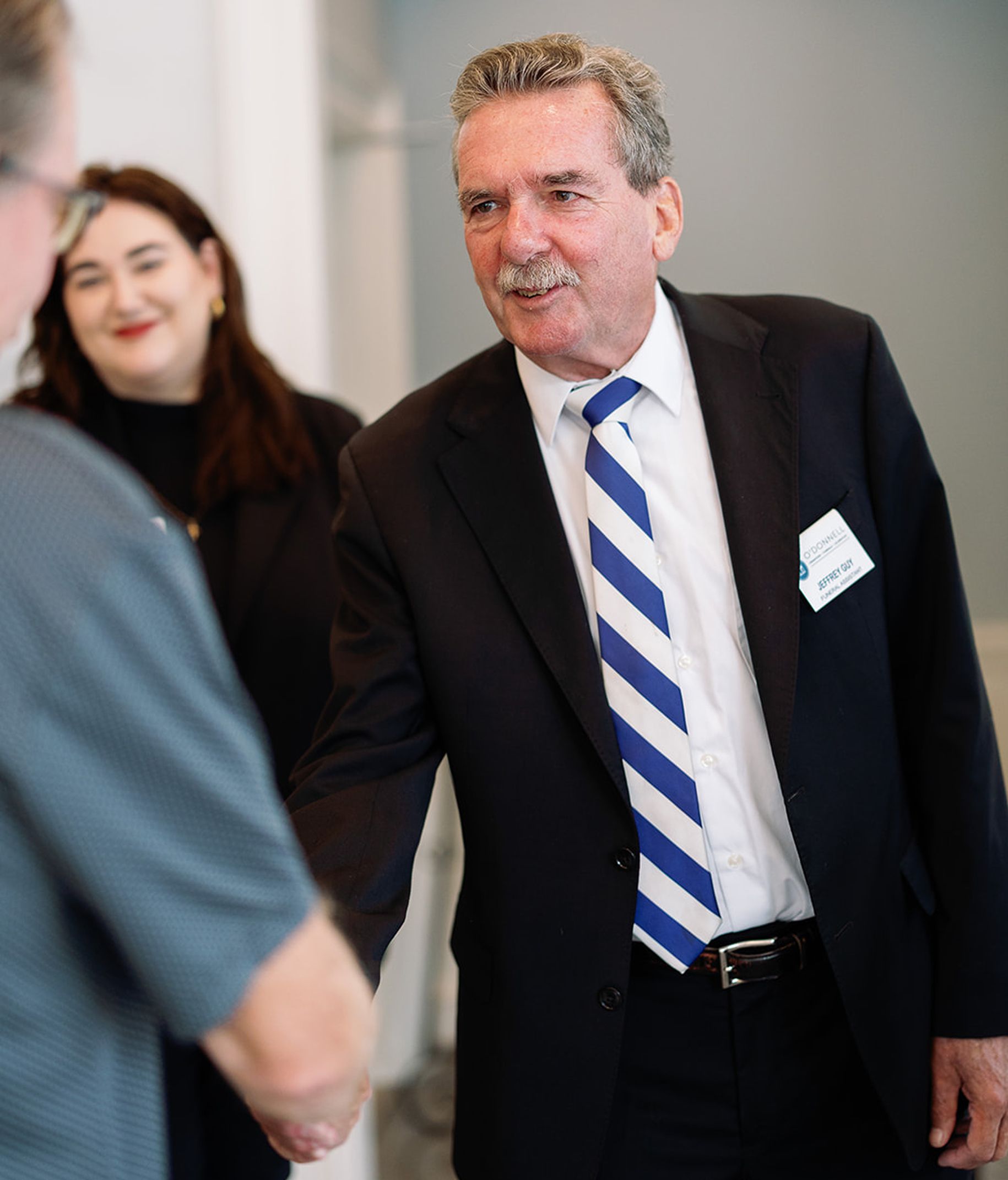 Man in suit shakes hands, smiles. Another person and woman with a friendly face are visible.