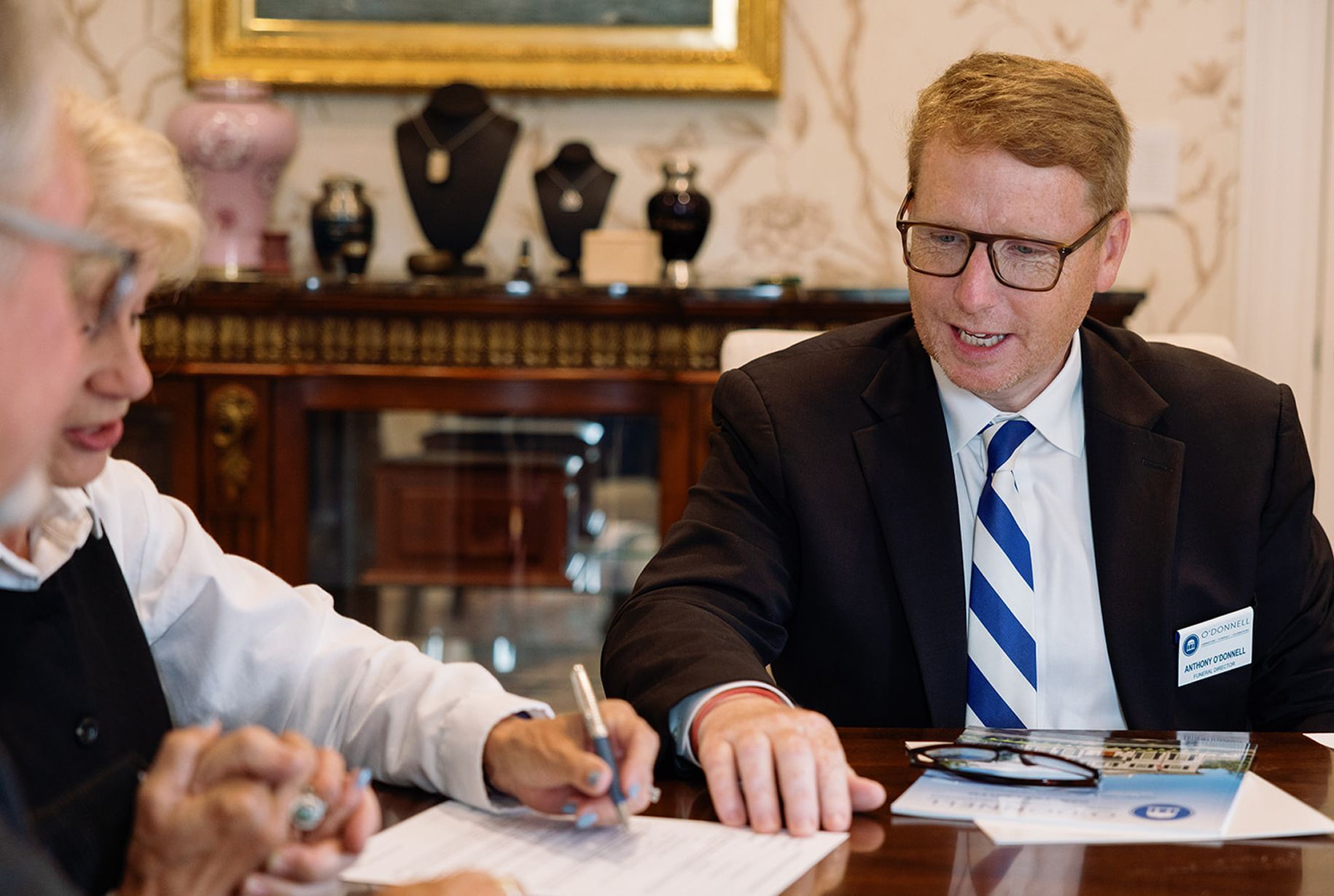 A man with glasses in a suit gestures as he speaks to two people seated at a table.