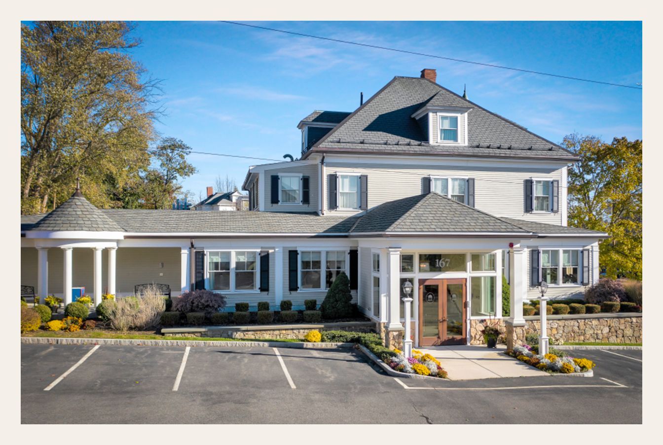 Gray building with a two-story main section and a single-story wing. There is a parking lot.