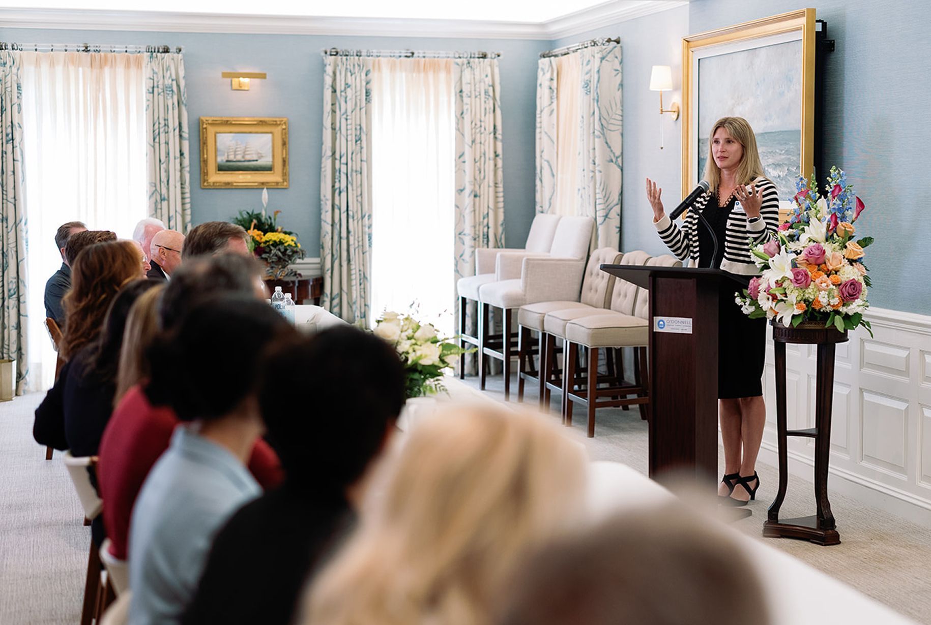 Woman speaking at a podium to an audience in a light blue room. Flowers, chairs, and windows are visible.
