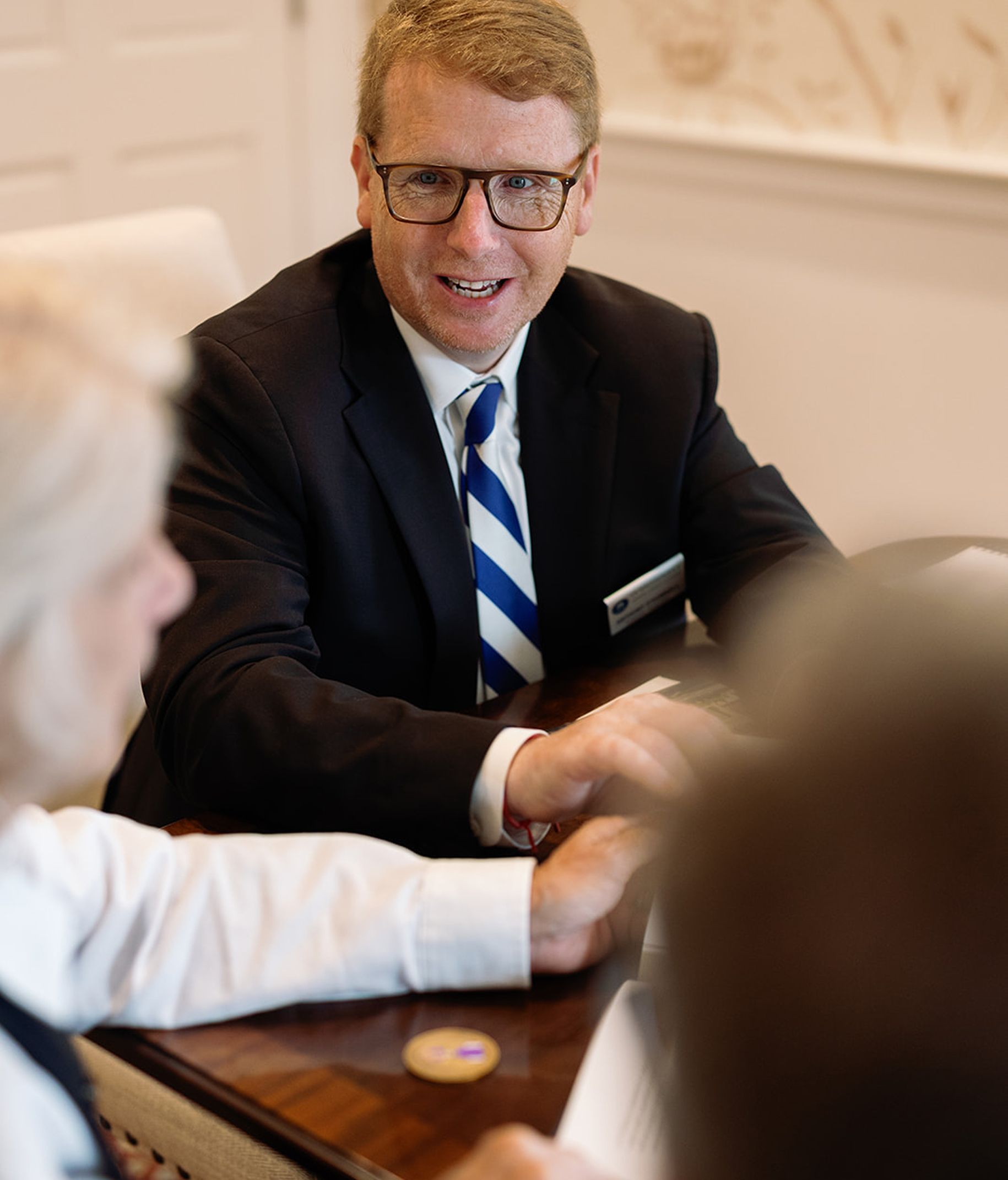 Man in suit and glasses smiles, seated at a table with others; blue tie.