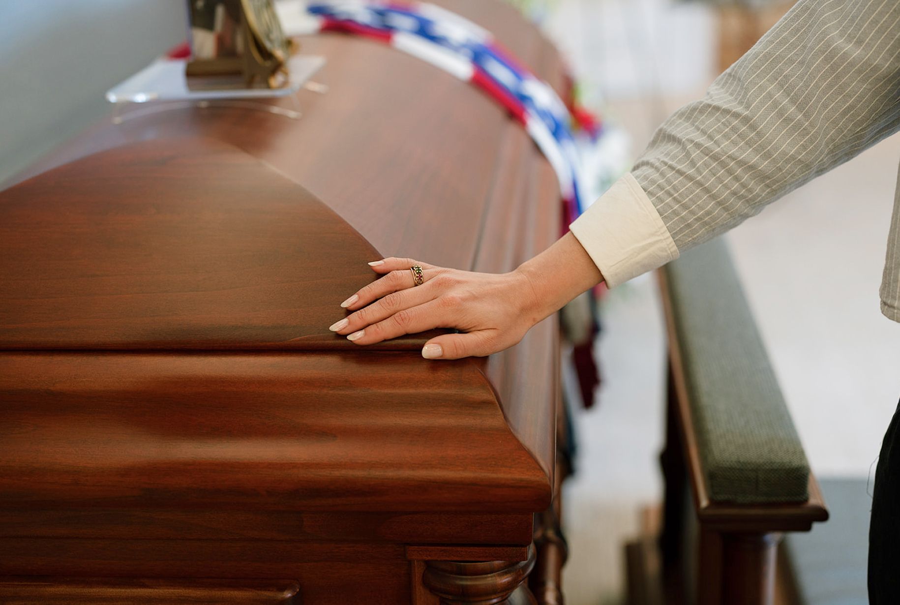 Person's hand resting on a polished wooden casket. A religious emblem and ribbon are on top, near a pew.