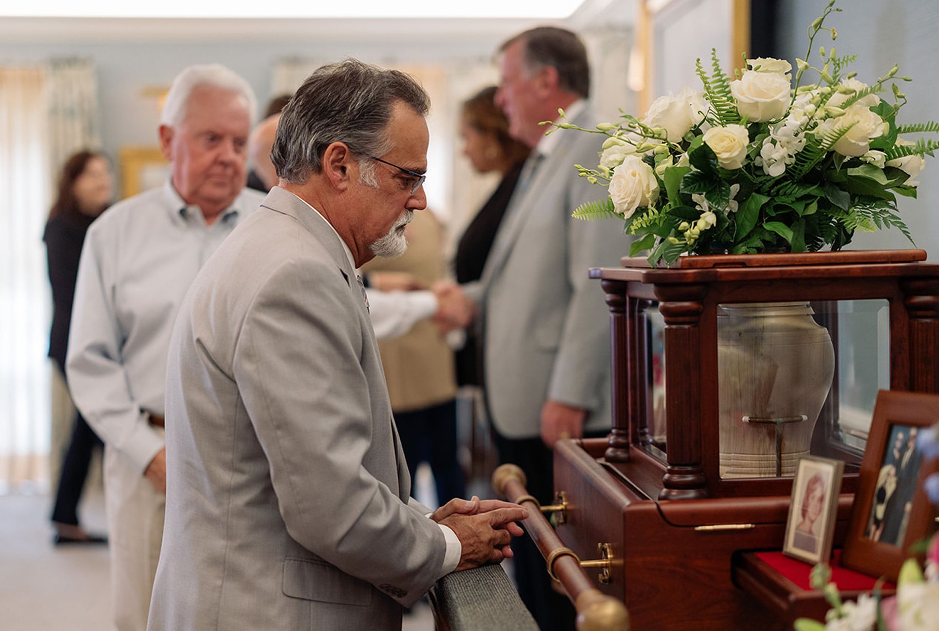 Mourners at a funeral, gazing at a casket. A floral arrangement sits atop.