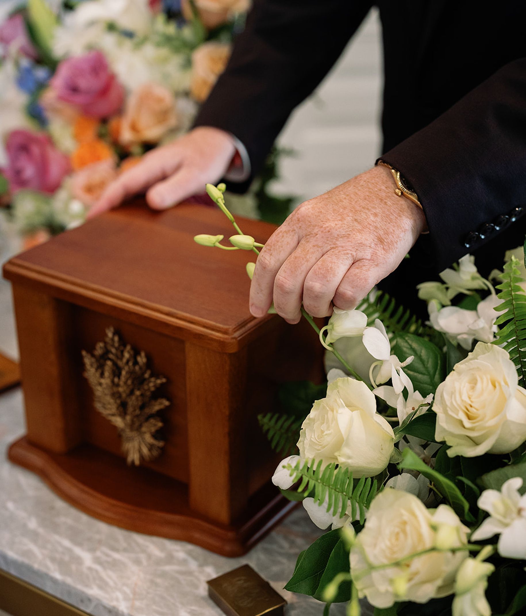 Person's hands with flower touching a wooden urn, surrounded by white roses and floral arrangements.