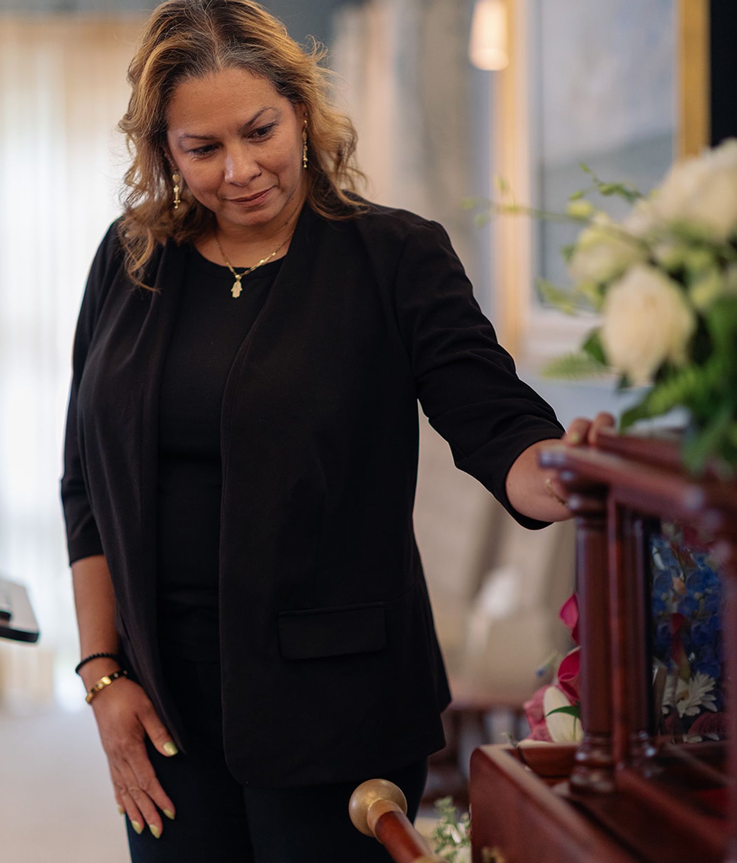 Woman in black blazer at a podium, looking down. Interior with flowers and window.