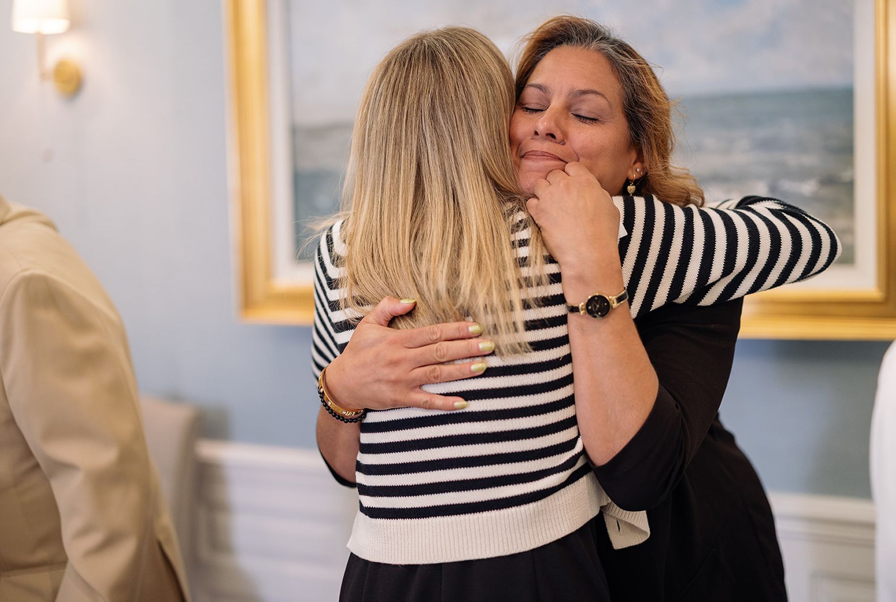 Two women embrace indoors, one in black and white stripes, the other in black, both smiling.