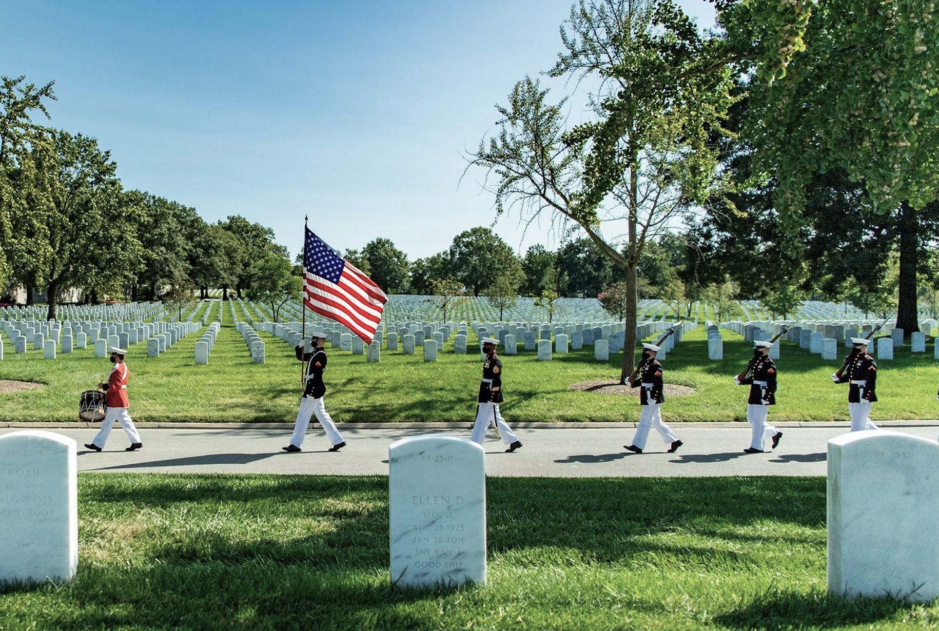 Soldiers marching in formation, carrying US flag, through a cemetery lined with white headstones on a sunny day.