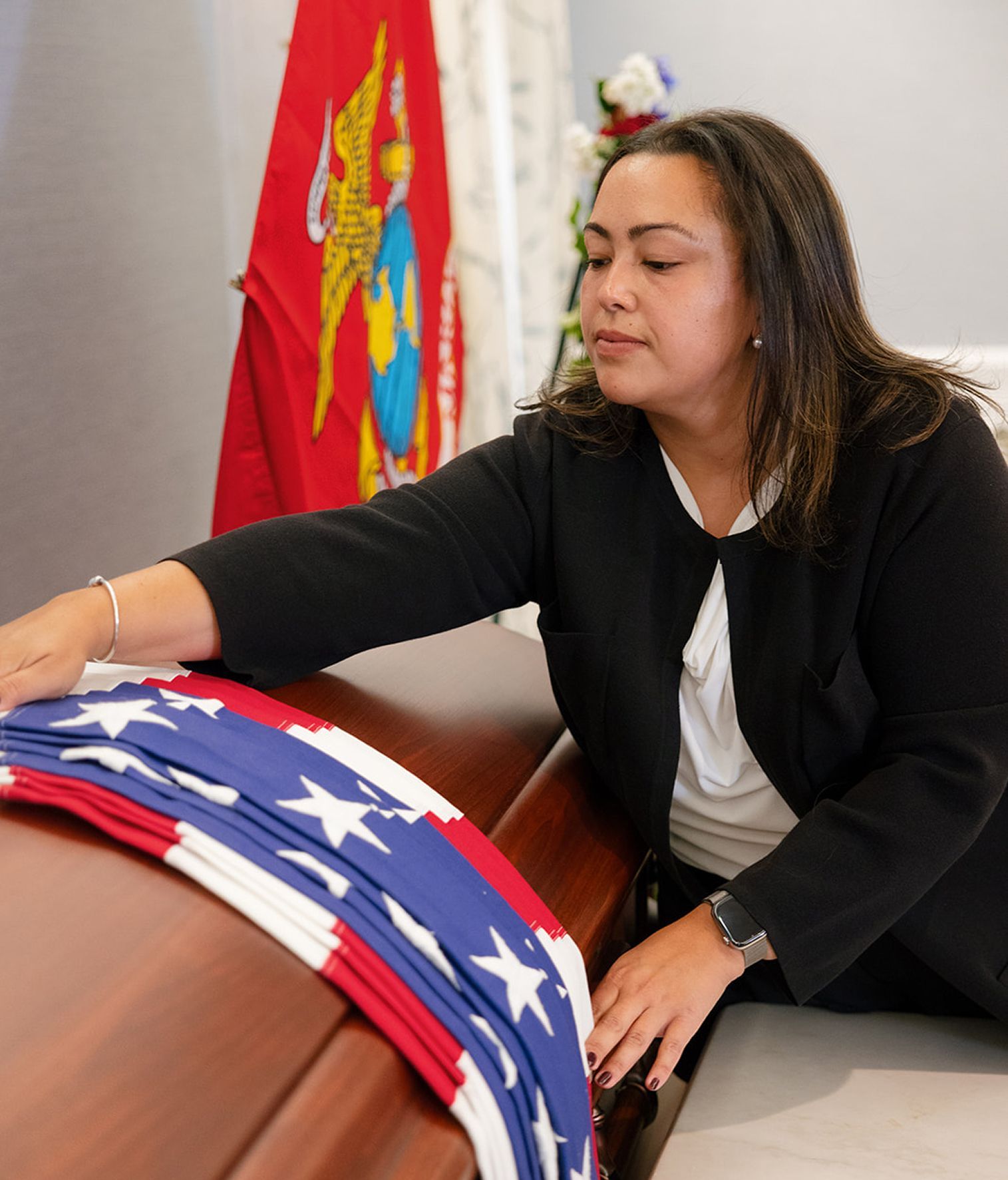 Woman in black blazer carefully folds a U.S. flag over a wooden coffin, Marine flag in the background.