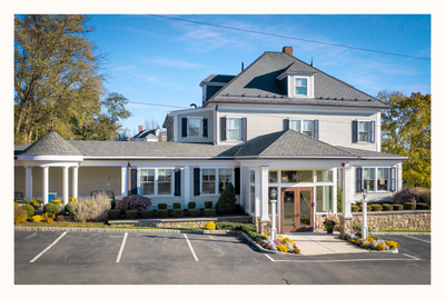 Light grey two-story building with multiple windows, black shutters, and parking in front, under a sunny sky.