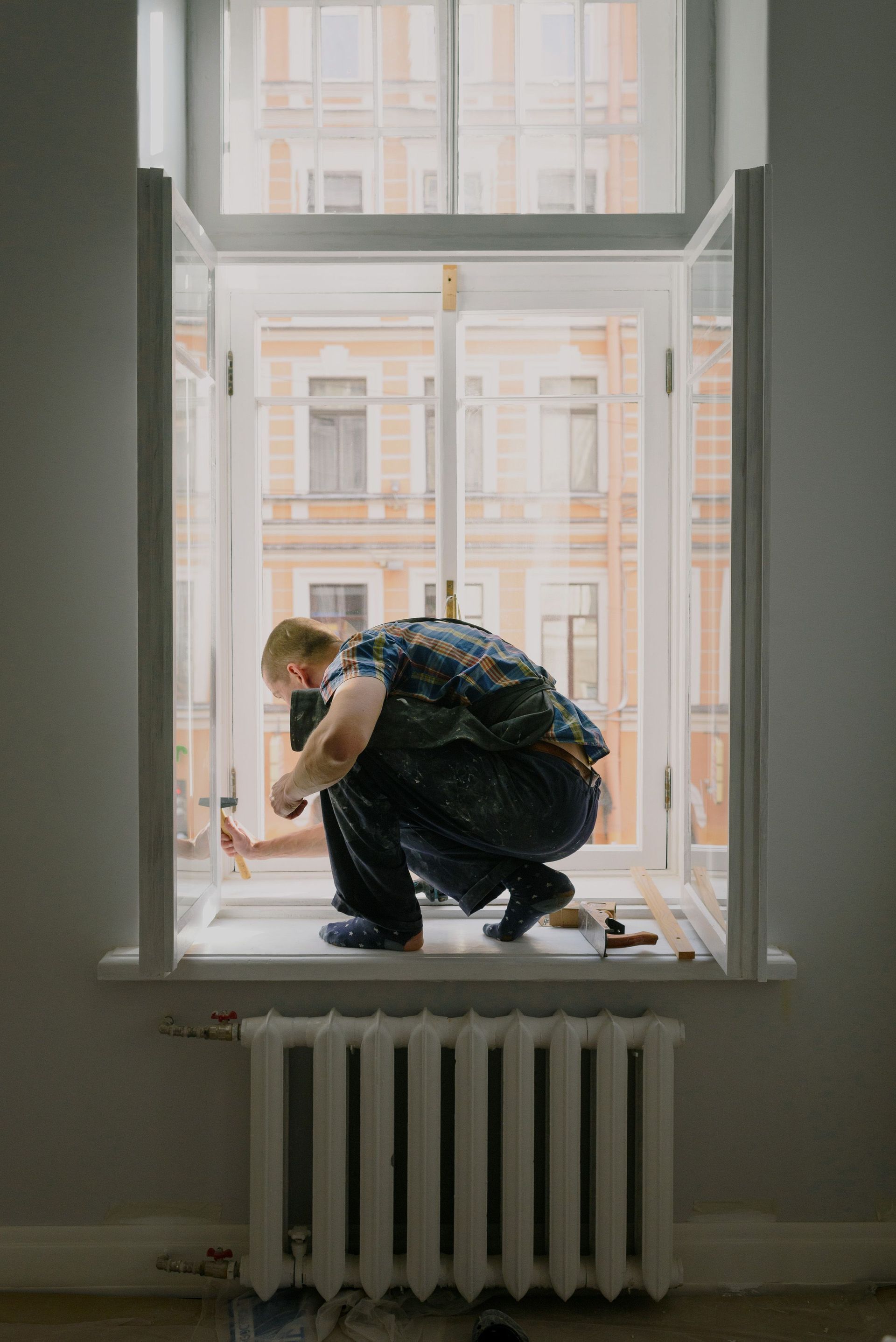 Person crouching on a windowsill in a room, looking outside at a building across the street