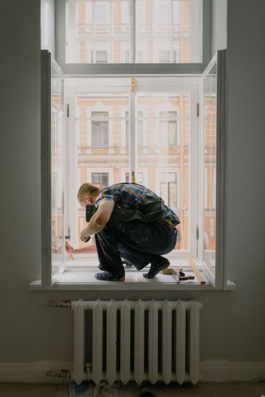 Person crouching on a windowsill in a room, looking outside at a building across the street