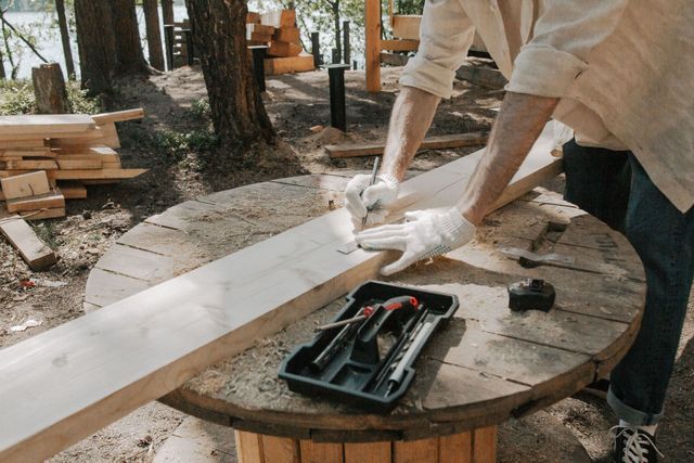 Person measuring a long wooden plank on a round outdoor table with tools nearby