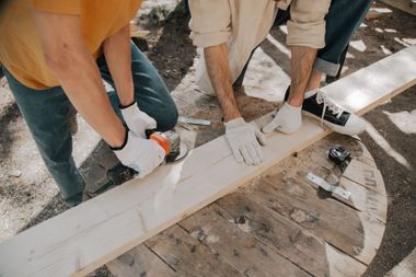 Three people in gloves painting a long white wooden beam on a worksite.