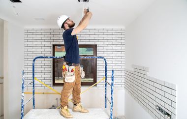 Worker installing ceiling fixture in a white-tiled room on a scaffold.