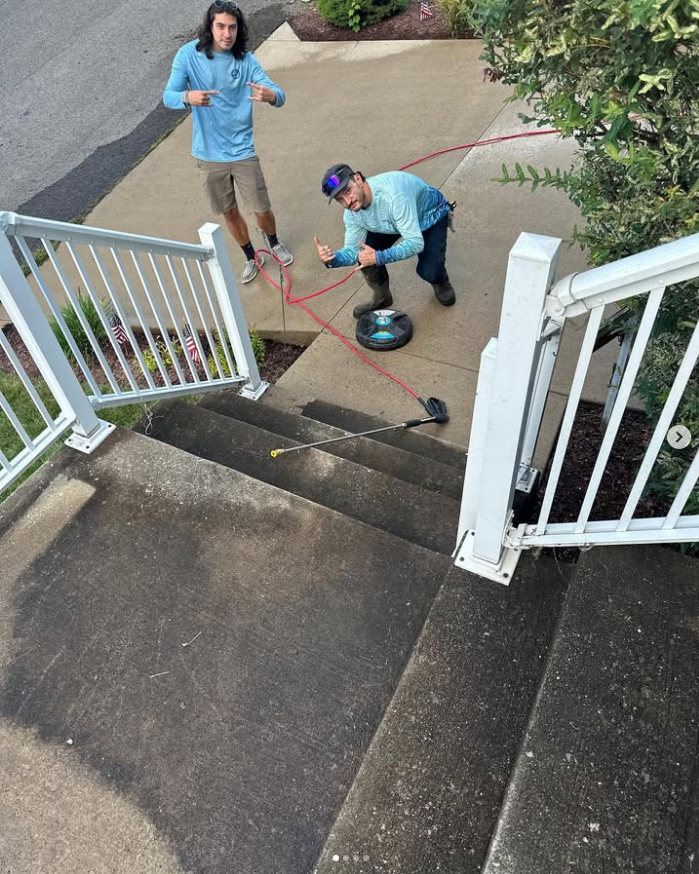Two people clean a wet concrete entryway with a pressure washer and hose.