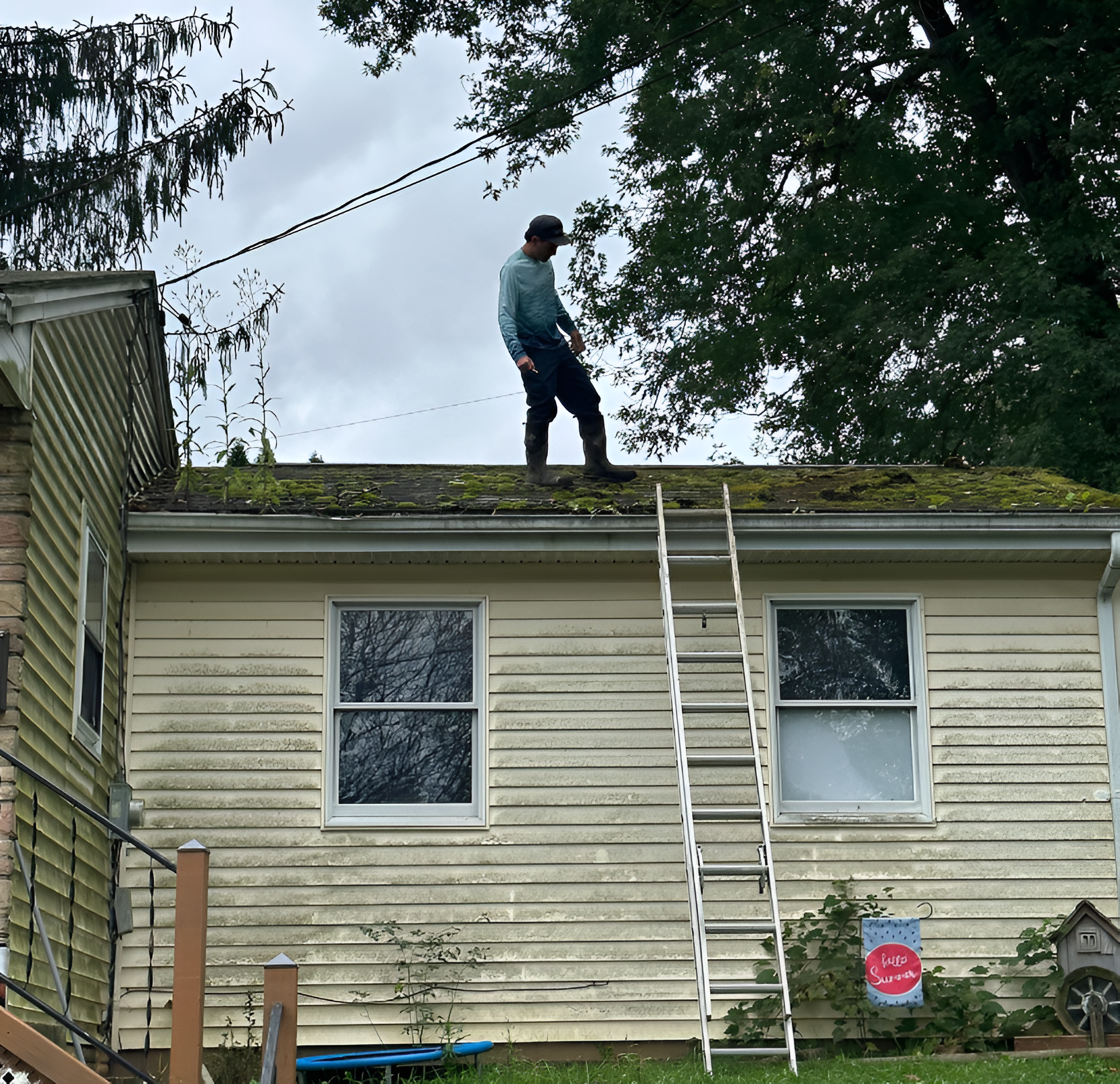 Person standing on a roof next to a ladder beside a beige house with moss on the shingles