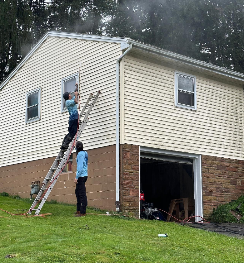 Two people on ladders painting the upper wall of a beige house beside an open garage.