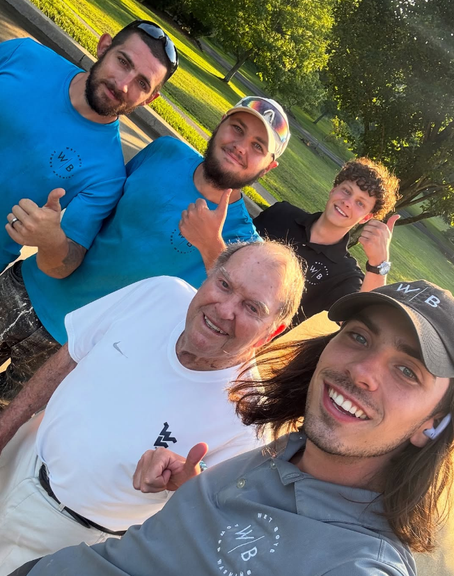 Five people smiling for a selfie outdoors in a sunny park, wearing casual shirts and caps.