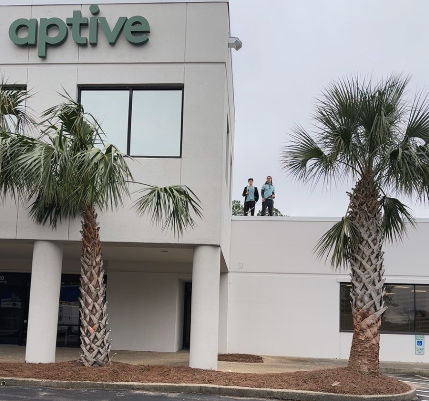 Office building with Aptive sign, palm trees, and three people standing on the roof under a cloudy sky