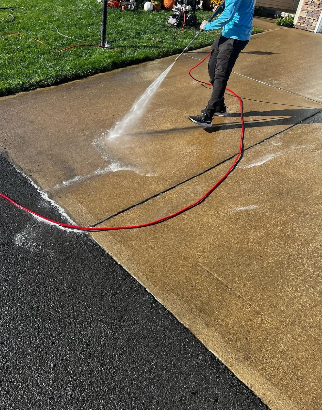 Person pressure-washing a concrete driveway with a red hose on a sunny day