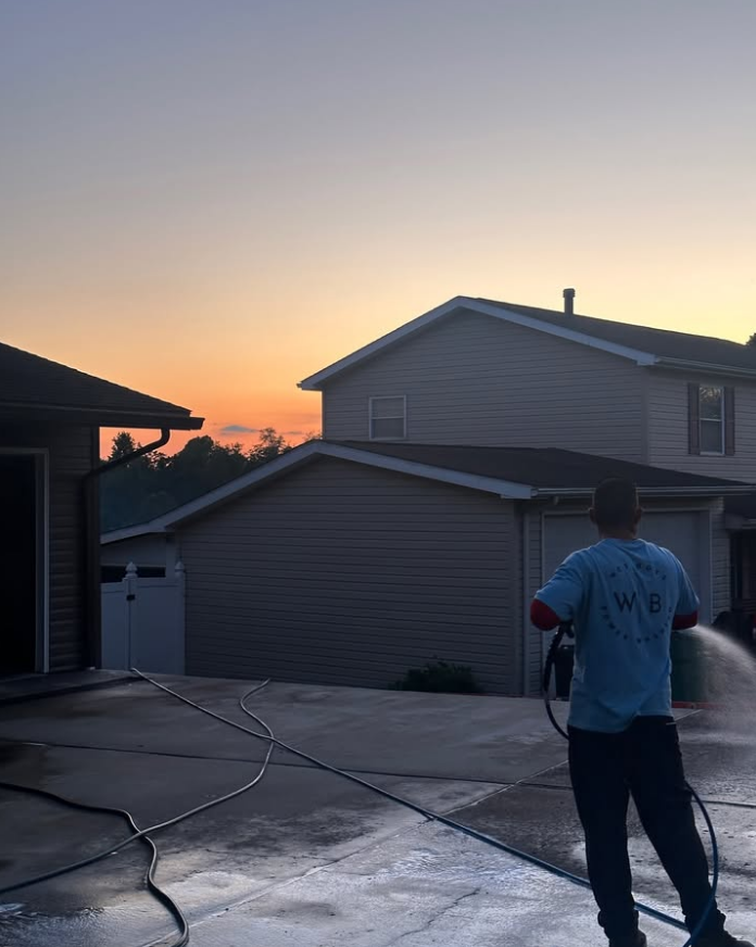 Person spraying water on a driveway at sunset, with houses and a hose in the background