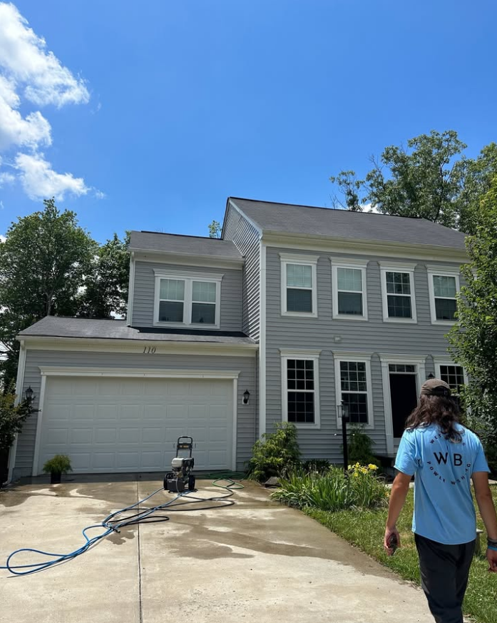 Two-story gray house with garage and driveway; a person stands in the foreground under a blue sky.