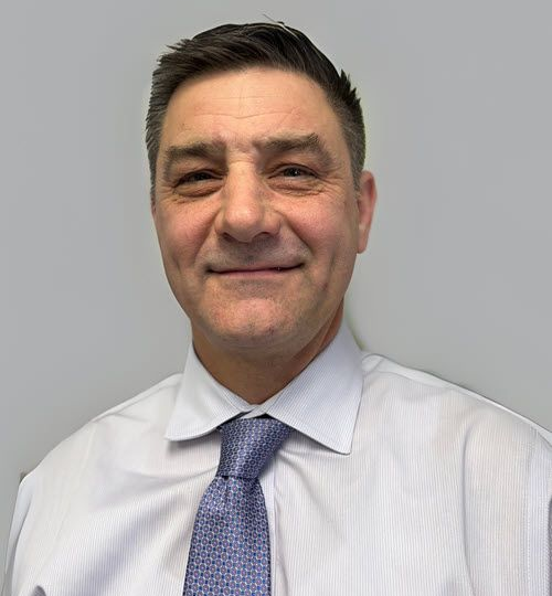 A headshot of a person with short dark hair, wearing a white dress shirt and a blue patterned tie against a grey background.
