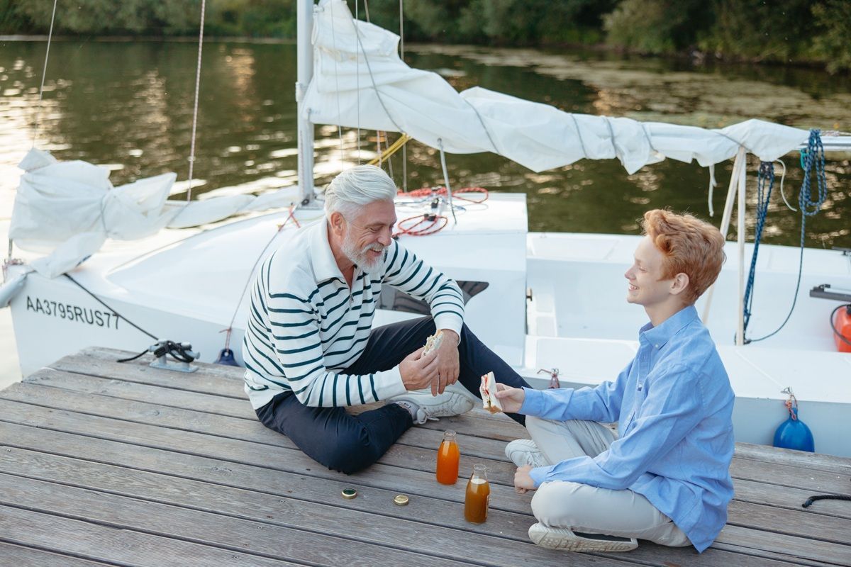 Two people sitting on a dock beside a small sailboat, talking and drinking by the water.