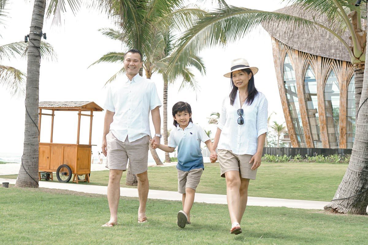 A family walks together on a green lawn by palm trees and a wooden building, smiling in matching light-colored outfits.