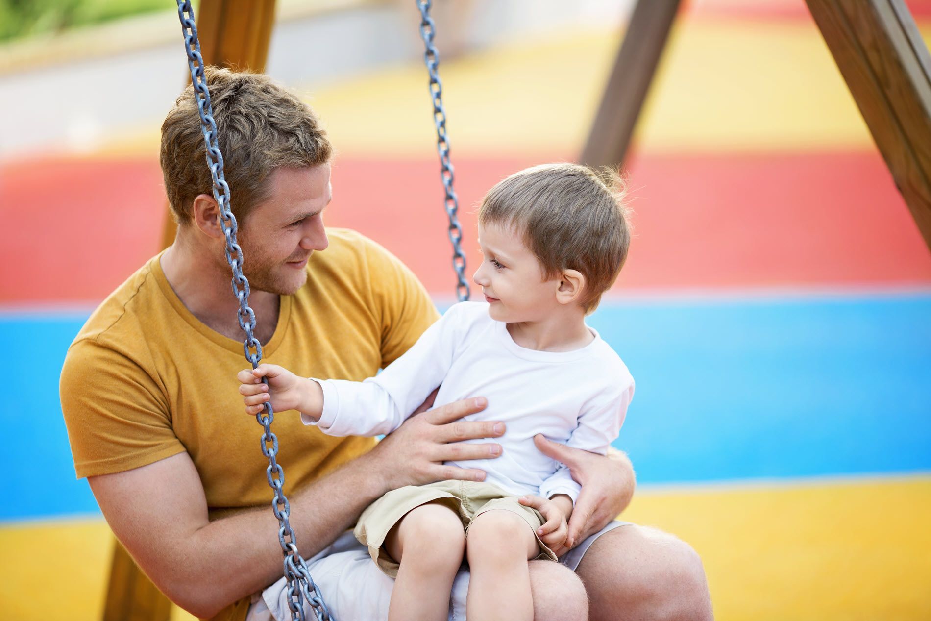 Father and child sitting on a swing, smiling at each other in a colorful playground.