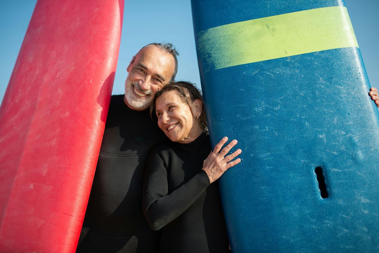 Couple in wetsuits smiling, holding surfboards: red and blue, outdoors, blue sky.