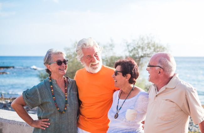 Group of four smiling seniors by the ocean, one wearing orange, all enjoying sunny day.
