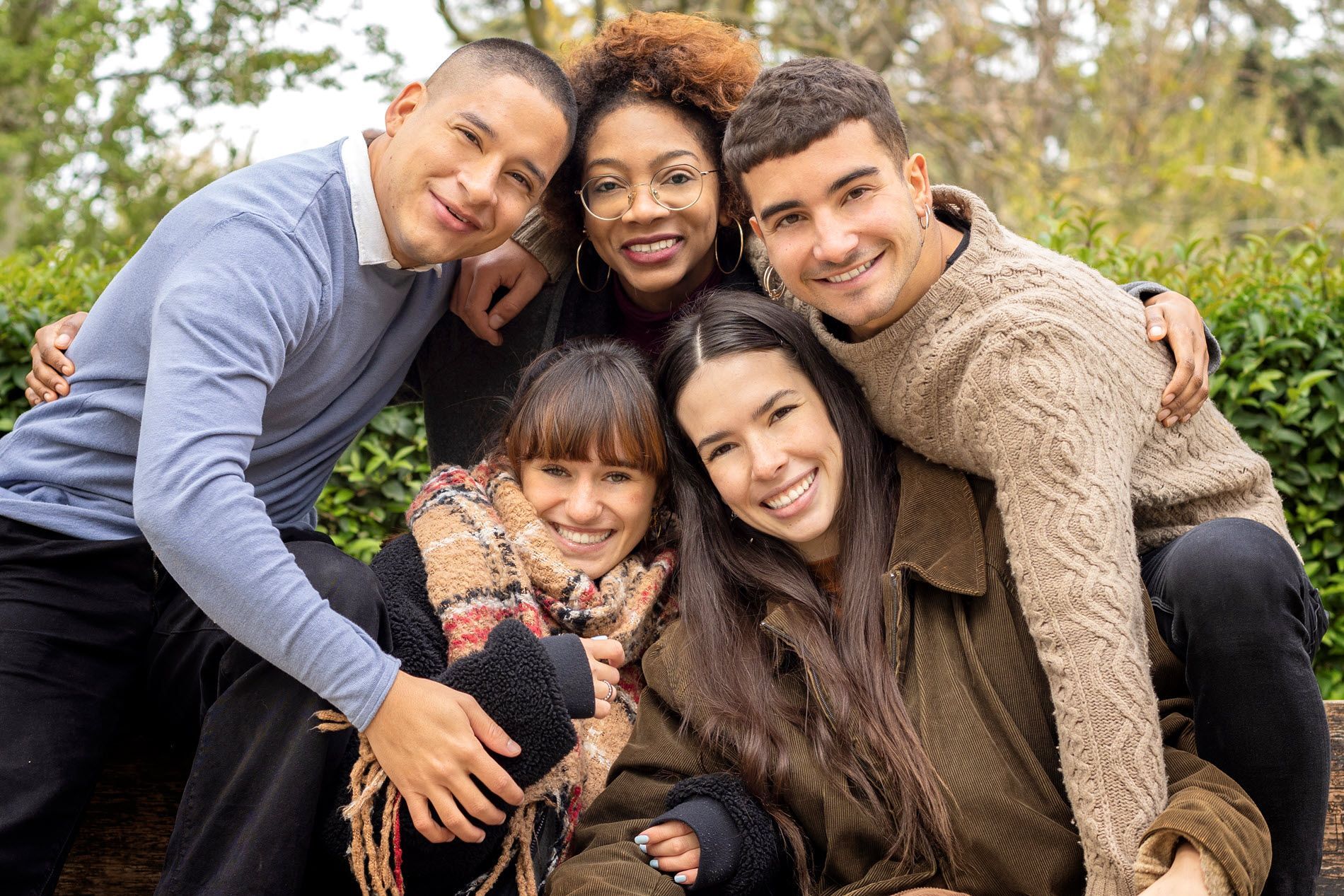 Group of five smiling friends huddled together outdoors, green background.