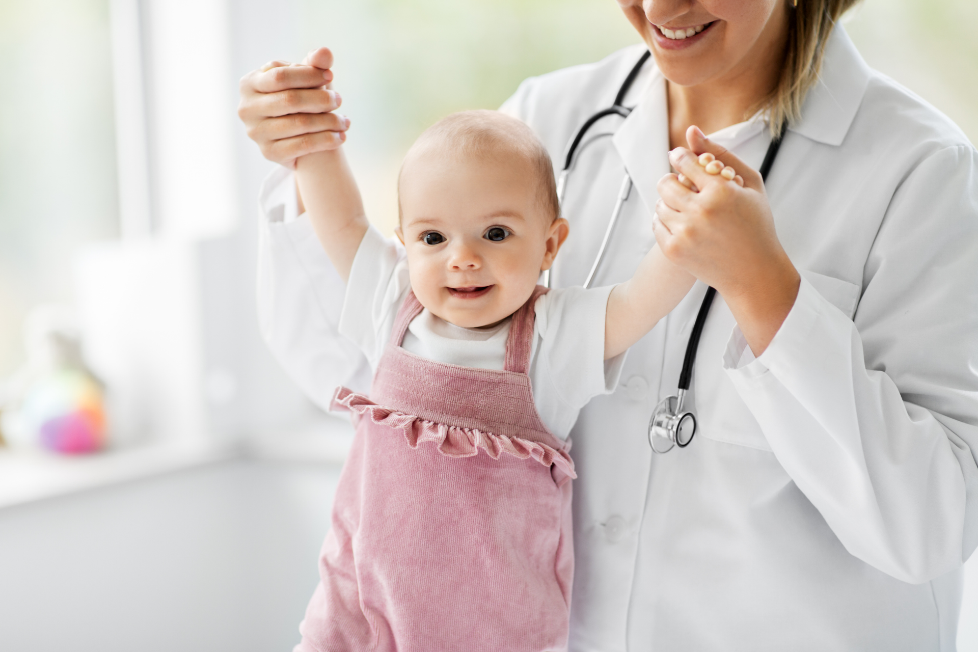 A children’s pediatrician supports a baby who is taking their first steps to assess for possible issues.