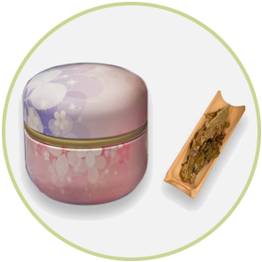 Pink and purple tea tin with floral design next to a bamboo tray holding green tea leaves.