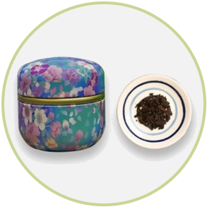 Floral tea tin next to a small plate with loose tea leaves; white, blue, teal, gold.