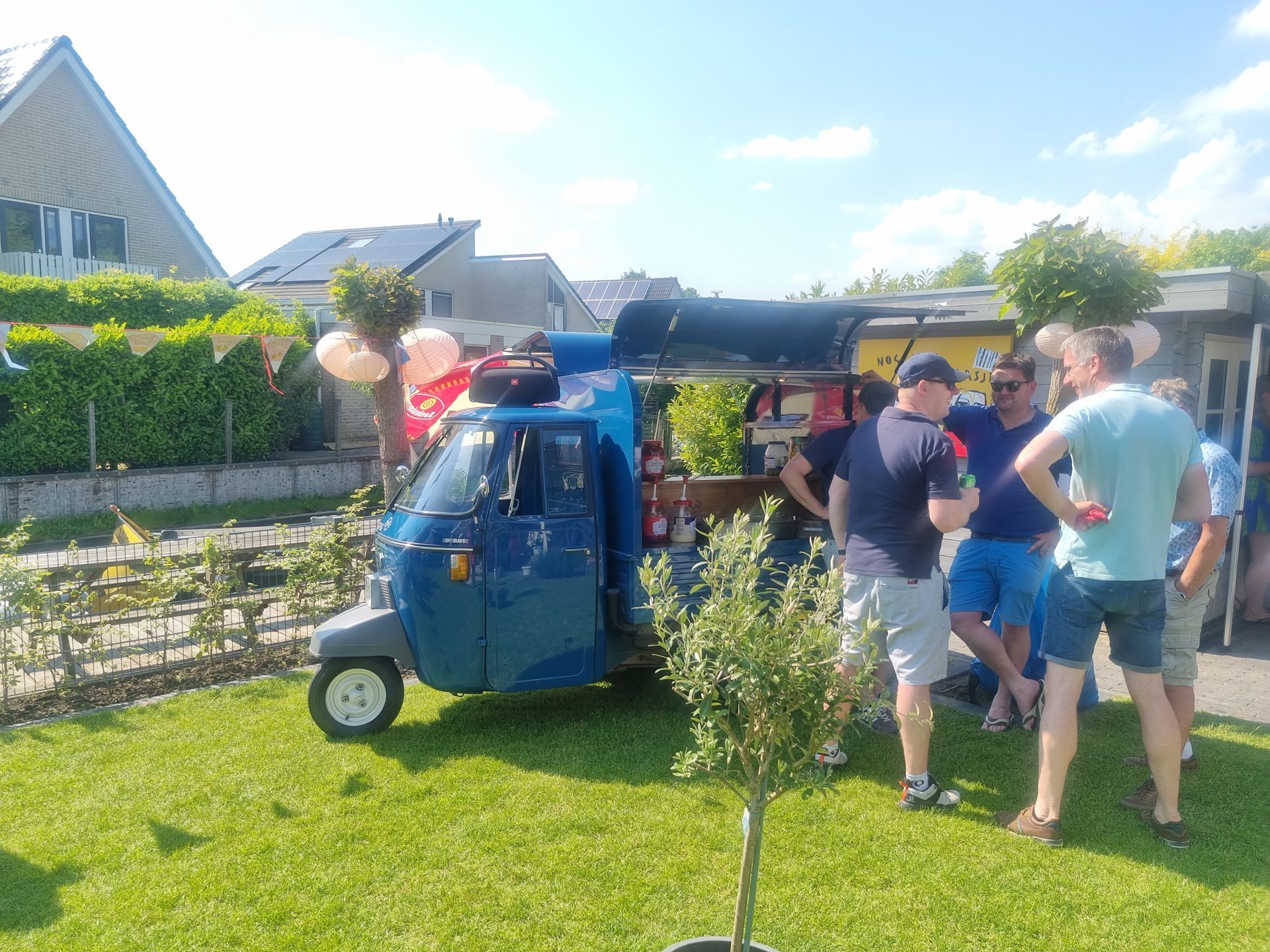 Een blauwe Piaggio Ape foodtruck met klanten op een grasveld op een zonnige dag.