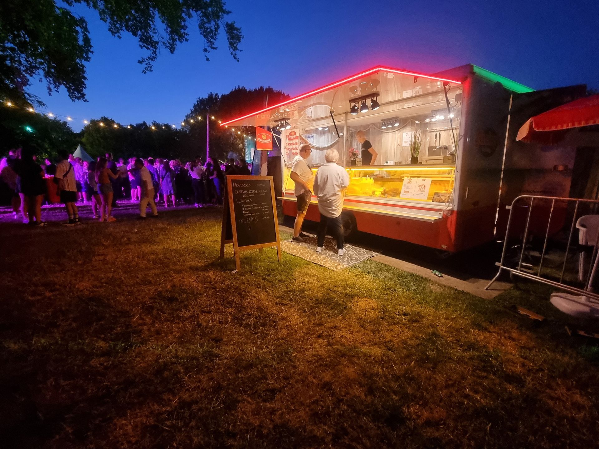 Een foodtruck 's nachts, verlicht met rode en witte lampen, bedient klanten. Mensen verzamelen zich in de buurt.