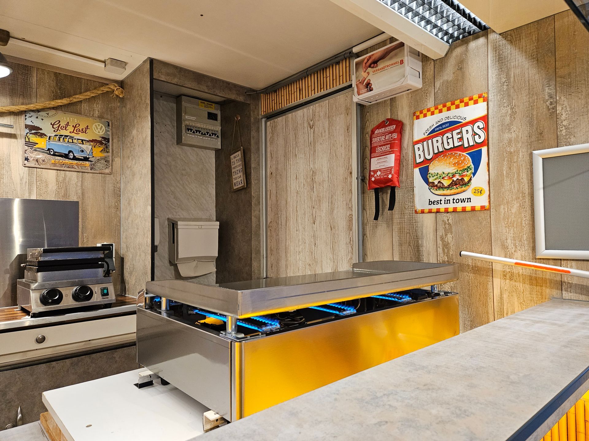 Interieur van een klein eethuis, met een toonbank, apparatuur en een hamburgerreclame aan de muur.
