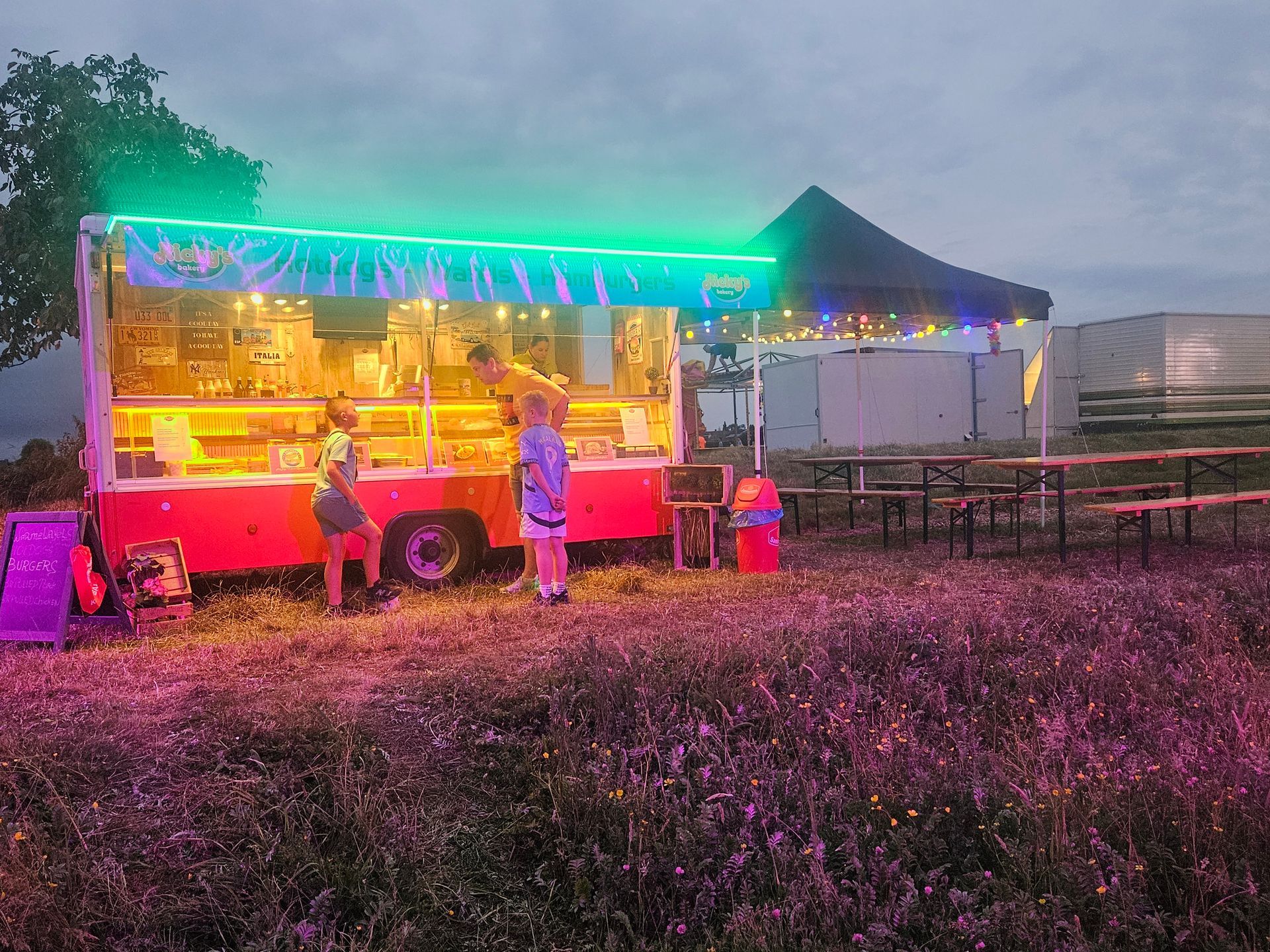Een foodtruck met neonverlichting en klanten buiten in de schemering.