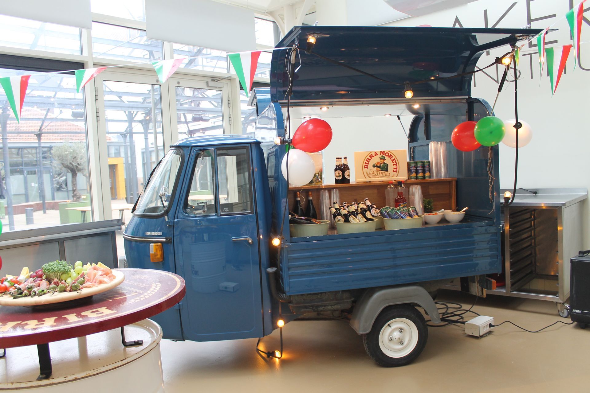 Blauwe foodtruck met open serveergedeelte; feestelijke versieringen; eten op de toonbank en op tafels.