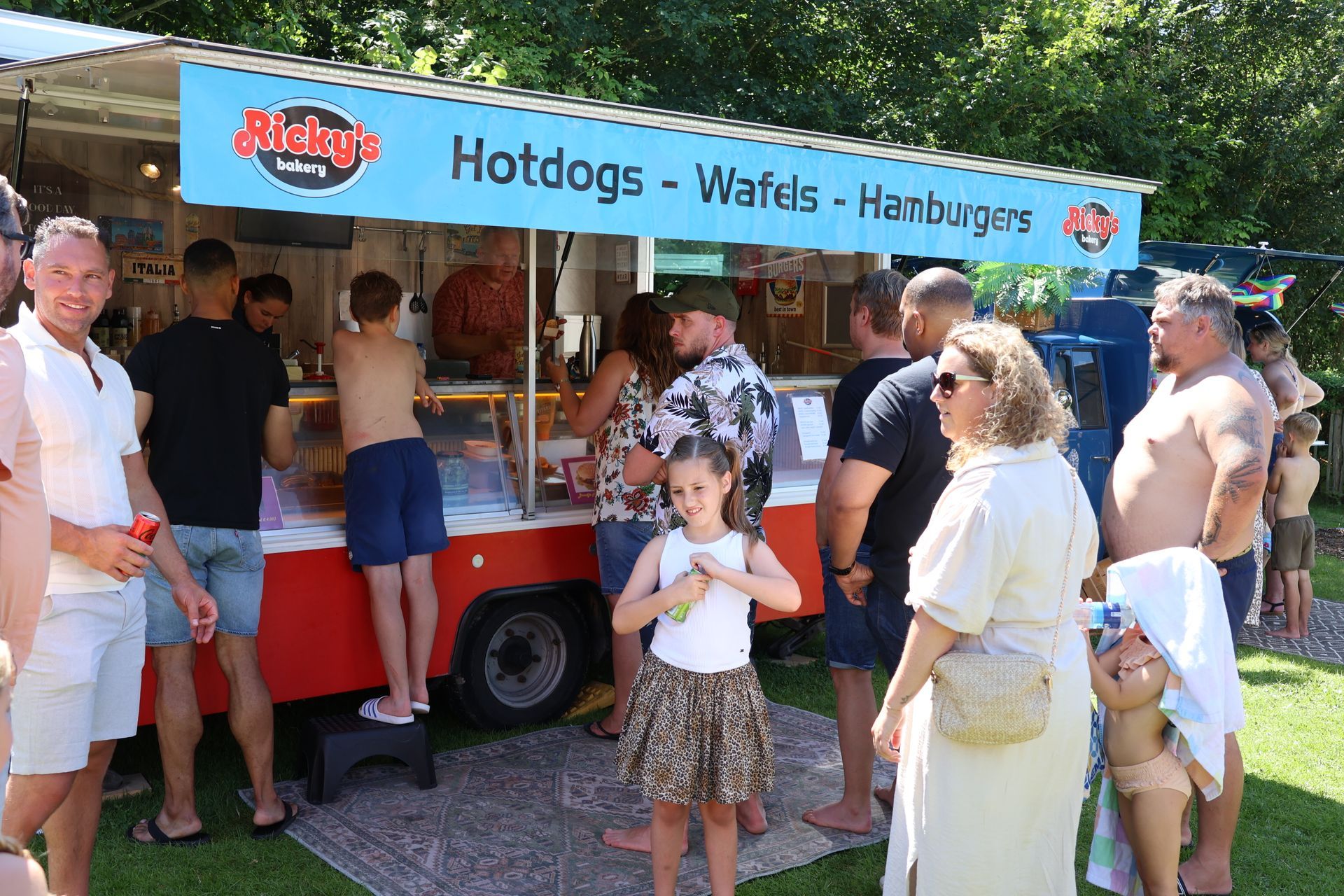 Mensen staan ​​in de rij bij een foodtruck die hotdogs, wafels en hamburgers verkoopt, buiten op een zonnige dag.