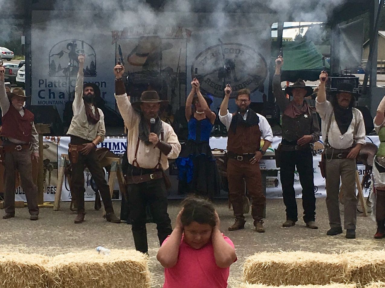 A girl in a pink shirt is covering her ears in front of a group of men dressed as cowboys.