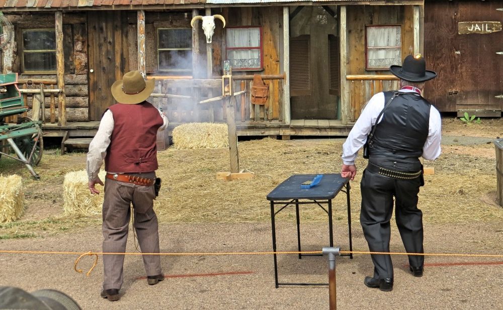 Two men in cowboy costumes are standing in front of a wooden building.