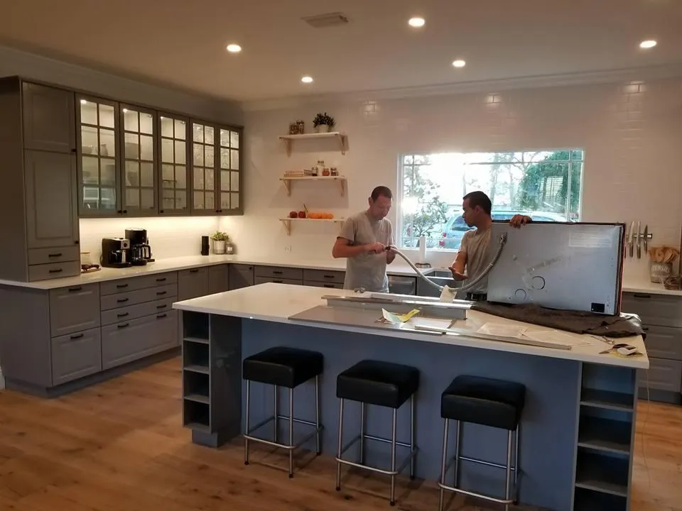 Two men are standing in a kitchen looking at a flat screen tv.