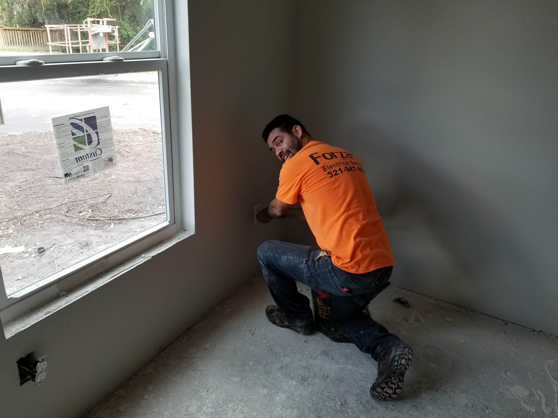 A man in an orange shirt is kneeling in a room next to a window.
