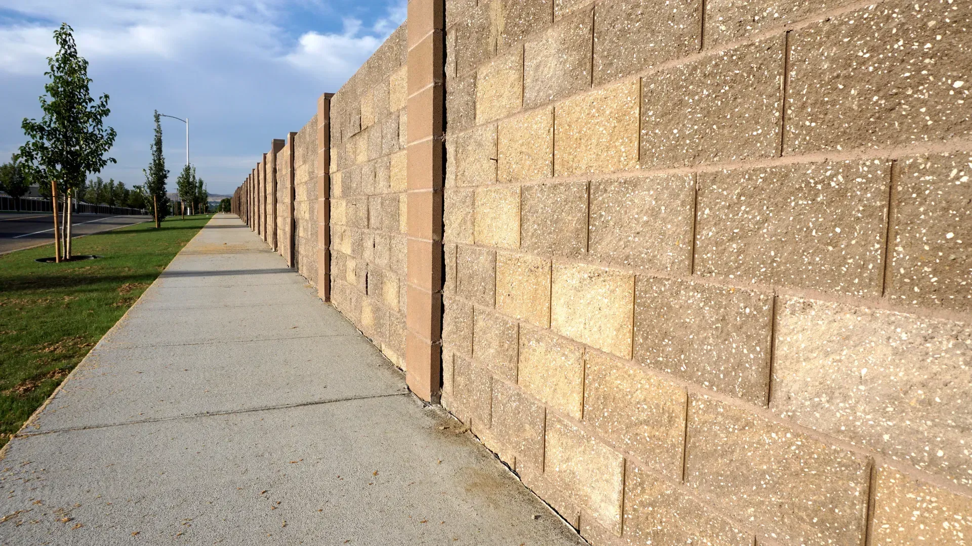 Sidewalk next to a brown concrete block wall, with grass and trees in the background under a blue sky.