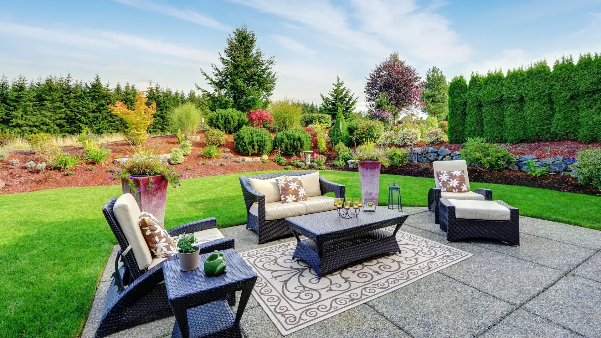 Outdoor patio with wicker furniture on a stone patio, lush green lawn, and colorful garden beds.