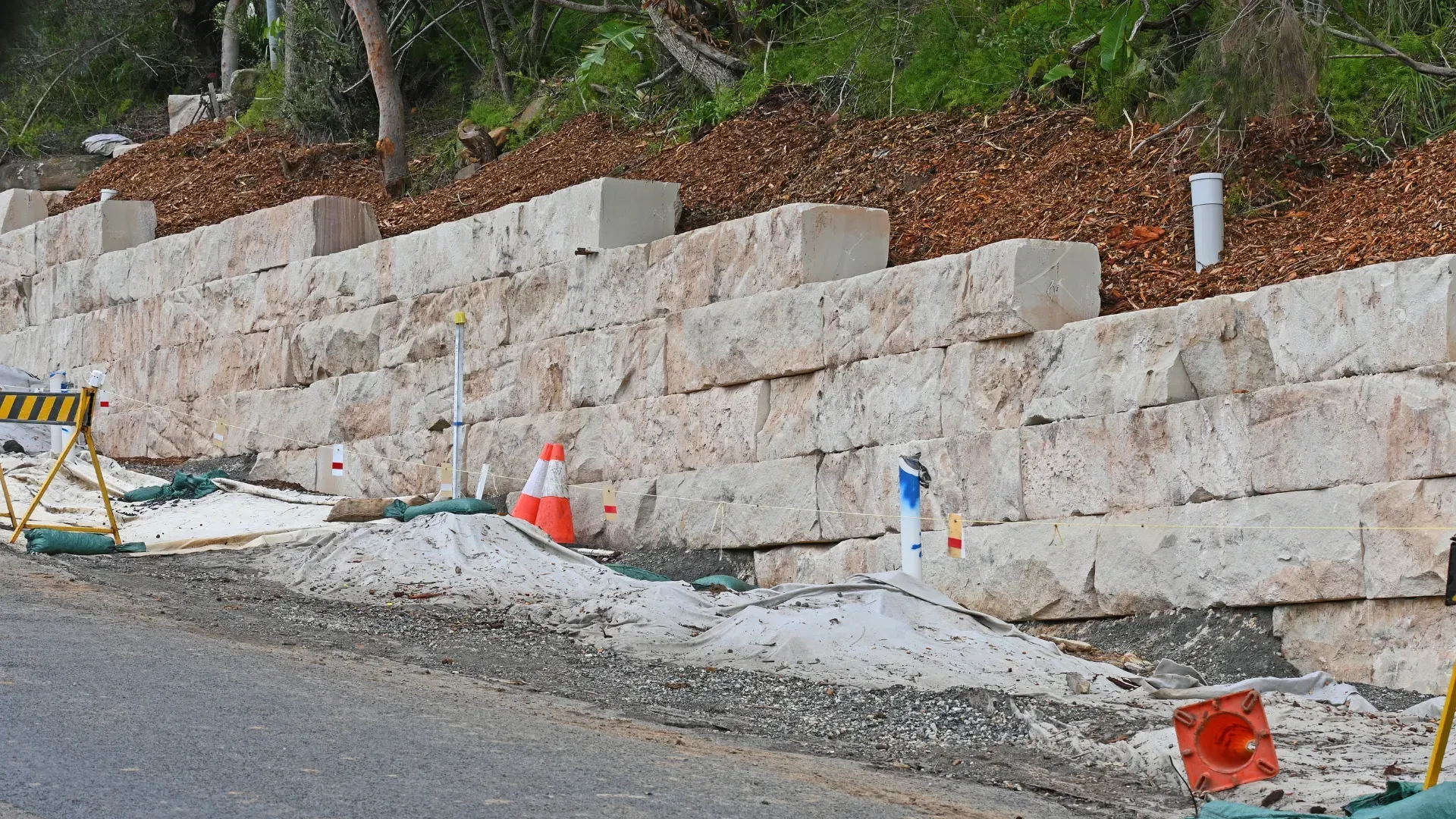 Stone retaining wall along a road with construction materials, safety cones, and a hillside in the background.